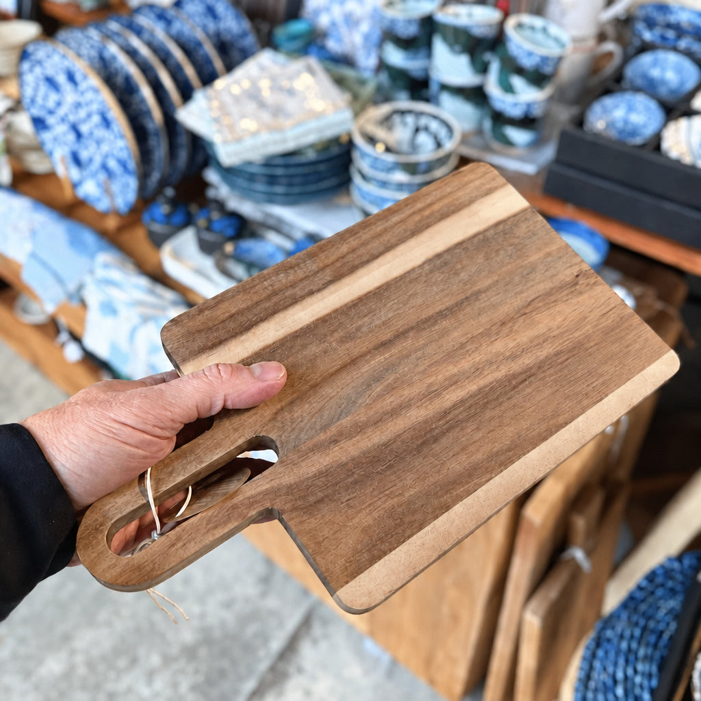 A hand holds a rectangular wooden cutting board with a handle, showing its natural wood grain and contrasting light wood edge, with a blurred shop background of ceramic dishes.