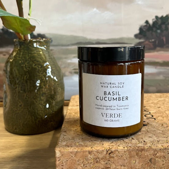 An amber jar candle labeled 'BASIL CUCUMBER' by Verde sits on a cork block on a wooden table, next to a green ceramic vase with leaves, with a landscape painting in the background.