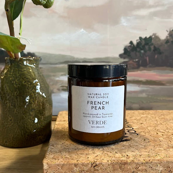 An amber candle jar labeled 'FRENCH PEAR' by VERDE sits on a cork block on a wooden table, next to a green ceramic vase with dried leaves, with a landscape painting in the background.