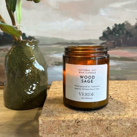 An amber jar candle labeled 'Wood Sage' by Verde sits on a cork block next to a green ceramic vase with dried greenery, set against a framed landscape painting.