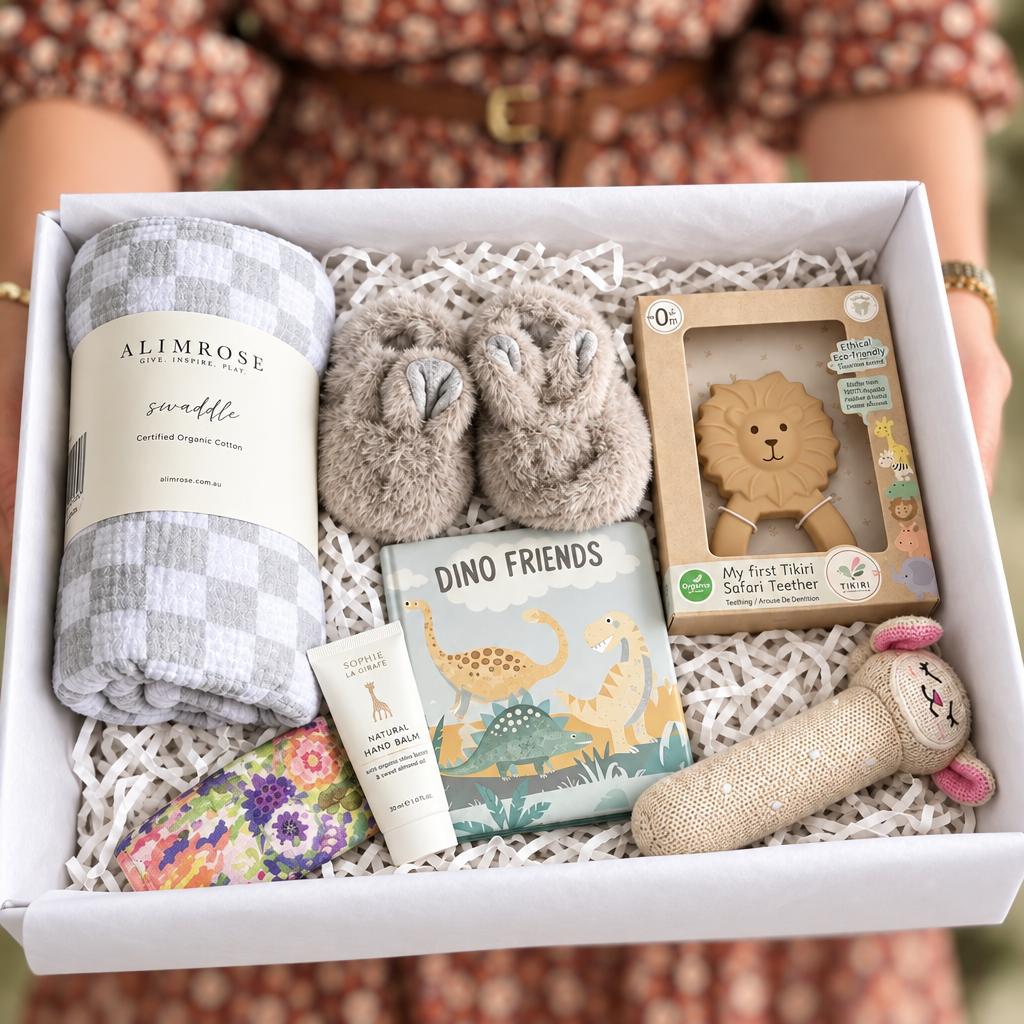 A curated baby gift box filled with baby items, including a gray and white checkered blanket, a pair of gray plush booties, a lion teething ring, a 'Dino Friends' book, a tube of lotion, and a floral sock, arranged in white tissue paper.