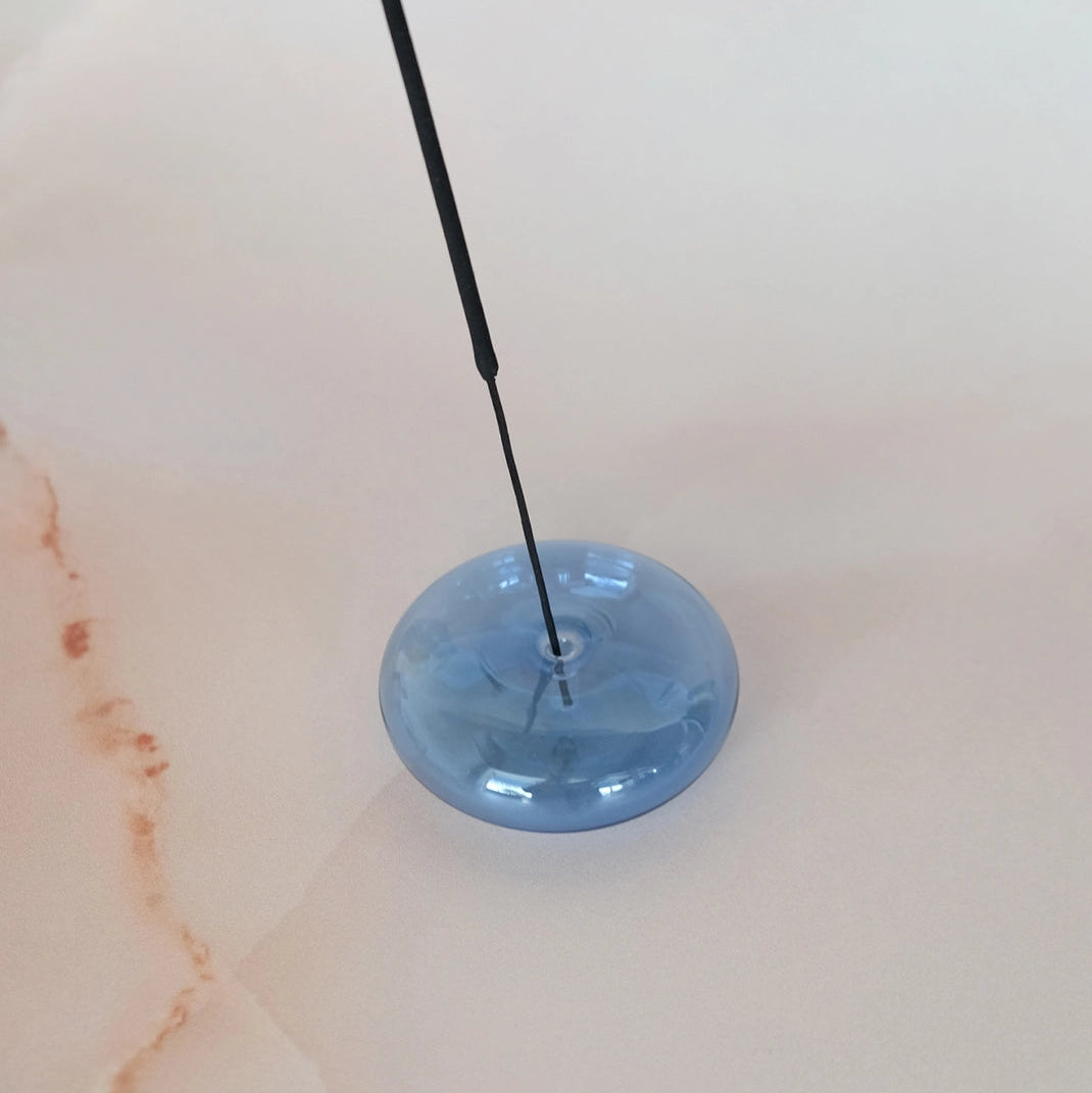 A light blue translucent circular glass dish with a black rod inserted into it, set against a warm off-white marble background.