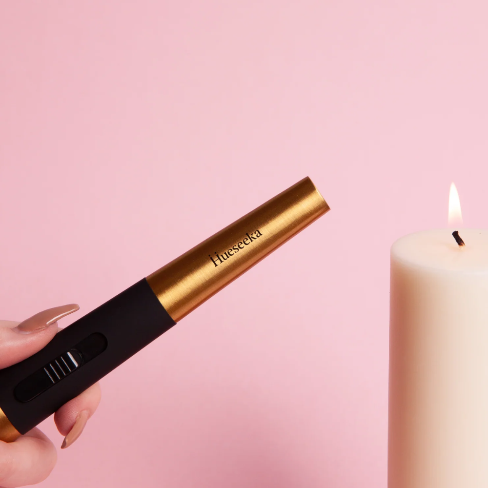 Hand holding a gold and black lighter next to a lit candle on a pink background