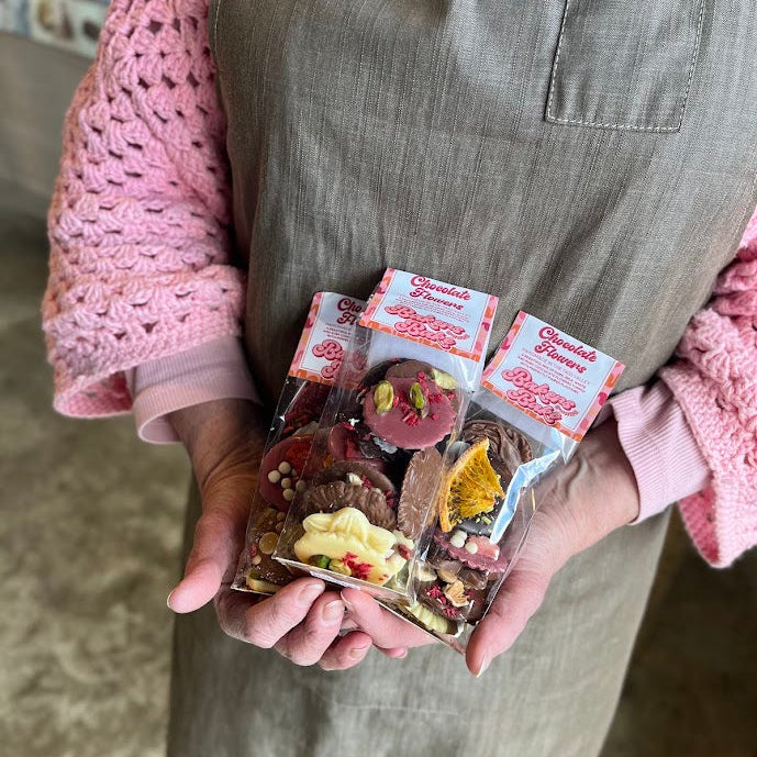 A person wearing a brown apron and pink knit sweater holds three small packaged gourmet chocolate treats in a clear plastic bag. The packages have white labels with red text, and the chocolates inside include pink, yellow, and dark chocolate pieces with decorative toppings.