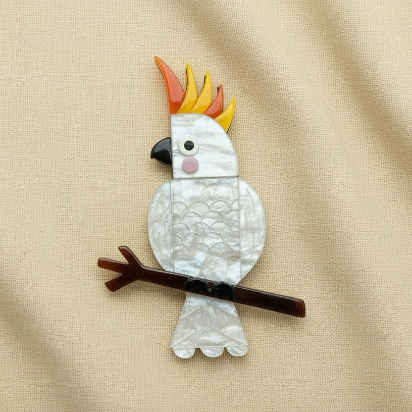 A cockatoo brooch made from acrylic, featuring a yellow and orange crested crown, a black beak, a pink cheek, and a brown branch perch, shown on a plain white background.