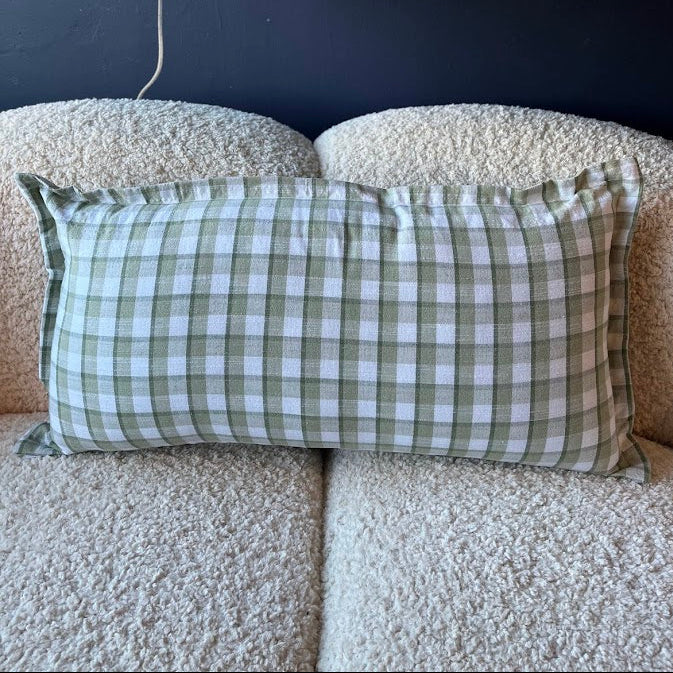 Green and white checkered pillow on a beige sofa with a dark wall and shelf in the background.