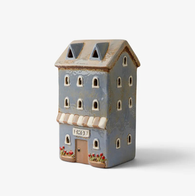 A decorative ceramic miniature house figurine in a distressed light blue-gray finish, featuring a brown door, cream awnings, arched windows, and small red flower details at the base, shown on a clean white background.