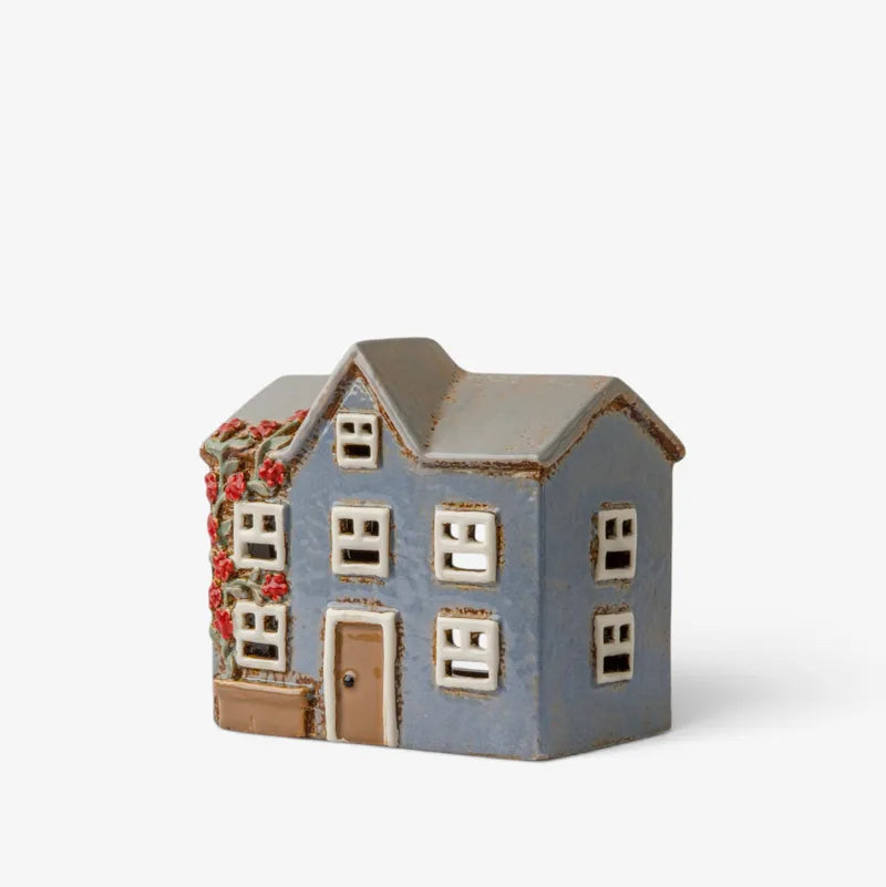 A small decorative house figurine with a blue-gray body, brown roof and door, white window frames, and red floral accents on the left side, photographed on a clean white background.