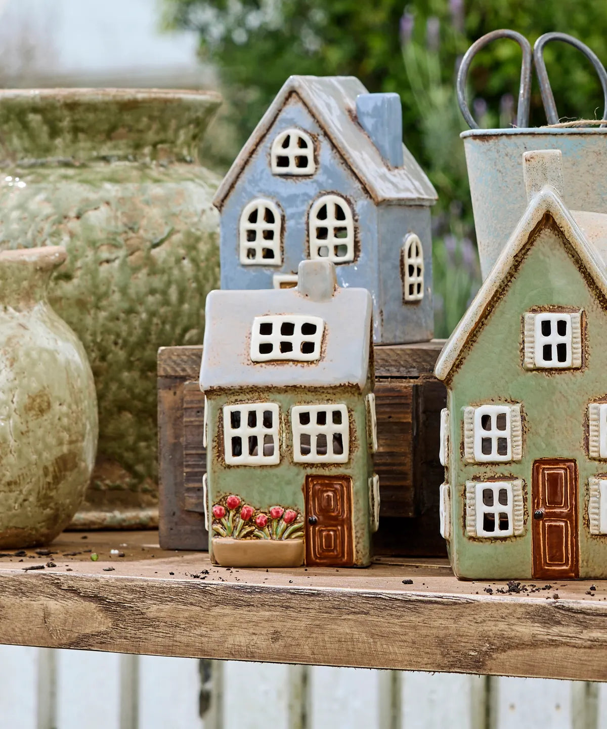 Three small ceramic house-shaped garden ornaments in pastel blue, sage green, and cream, displayed on a rustic wooden surface with other decorative vases in the background.