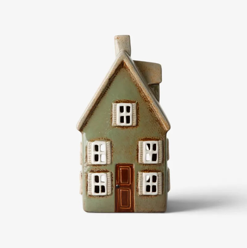 A small decorative house-shaped ceramic object with a muted sage green body, brown door and roof trim, and white window panes, photographed on a clean white background.