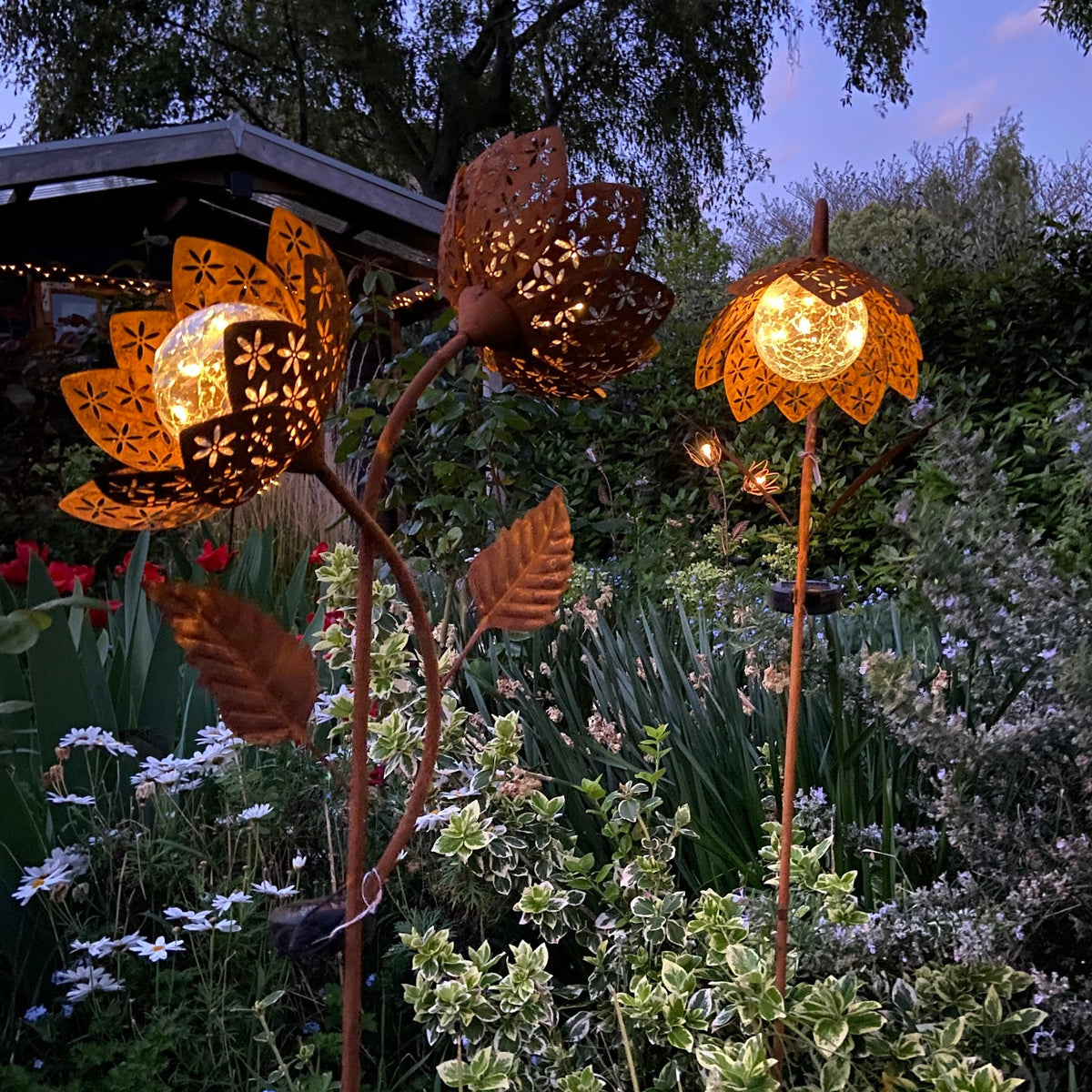 Three illuminated metal flower garden lights with cutout petals in a lush garden at dusk, casting warm light against green foliage and blue evening sky.