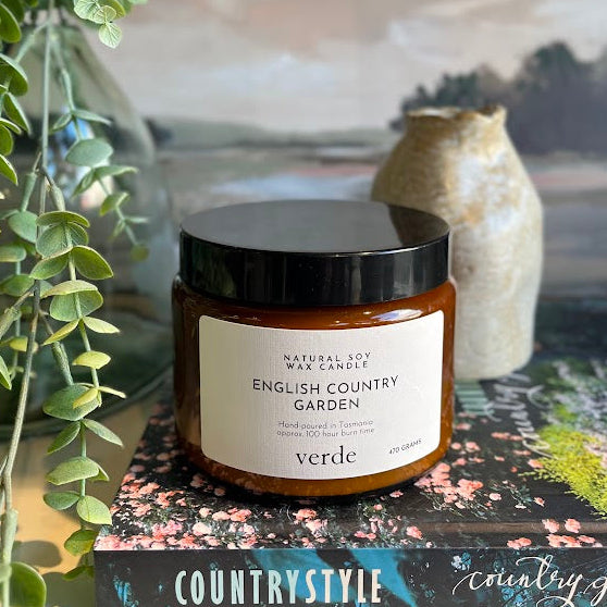 A jar candle labeled 'English Country Garden' by Verde sits on a book, styled with a green eucalyptus plant, a ceramic vase, and a framed landscape print in the background.