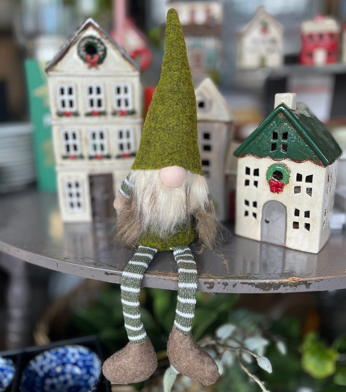 A festive Christmas gnome figurine with a green felt hat, white beard, striped green and white legs, and brown shoes, displayed on a shelf next to small ceramic houses.