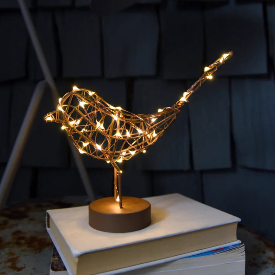 A decorative bird-shaped light fixture made of gold-toned wire with warm white fairy lights, mounted on a small round base and displayed on top of two stacked books against a dark textured wall.