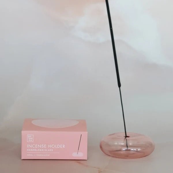 A pink glass incense holder with a black incense stick resting inside, shown next to its light pink packaging box on a light marble surface.