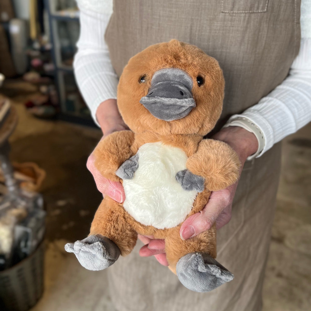 A small plush platypus toy being held in both hands by a person wearing a beige apron. The toy has a warm brown body, a white belly, and a grey beak, feet, and paws.