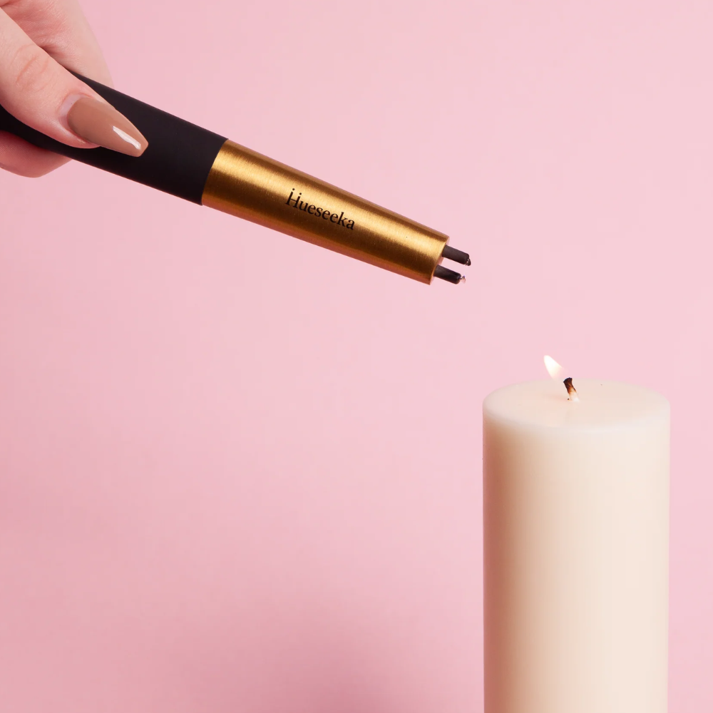Hand holding a gold lighter with black accents, towards a lit candle on a pink background.