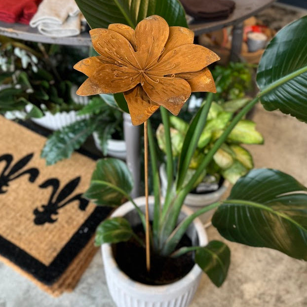 A decorative rusty flower garden stake with a warm brown flower and stem, inserted into a potted green plant in a white pot, set in what appears to be a retail shop environment.