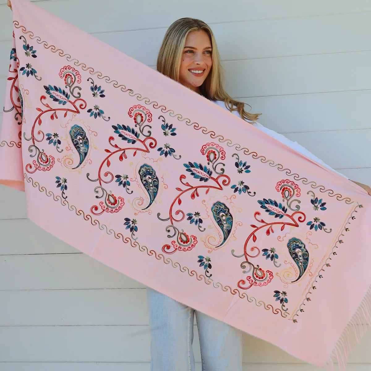 Woman holding a large pink scarf with floral and paisley patterns against a white wooden background.
