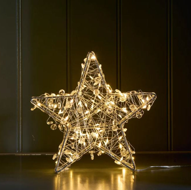 A glowing wire star decoration with warm white LED lights, standing on a reflective dark surface against a dark paneled background.
