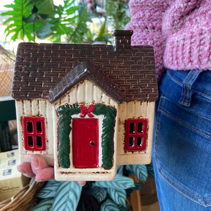 A ceramic Christmas house ornament or figurine with a brown shingled roof, cream siding, a red door, green wreath, and red window frames, held by a person wearing a pink sweater and blue jeans in a shop setting.