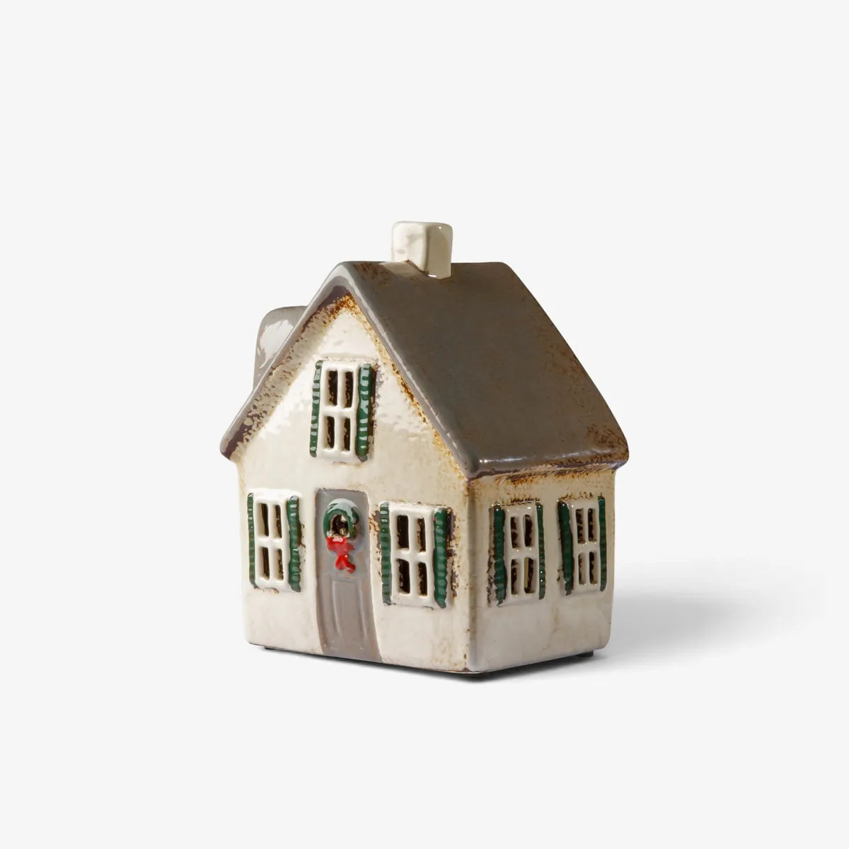 A small ceramic Christmas house figurine with a dark brown roof, cream-colored walls, green shutters, and a gray door with a wreath, set against a plain light gray background.
