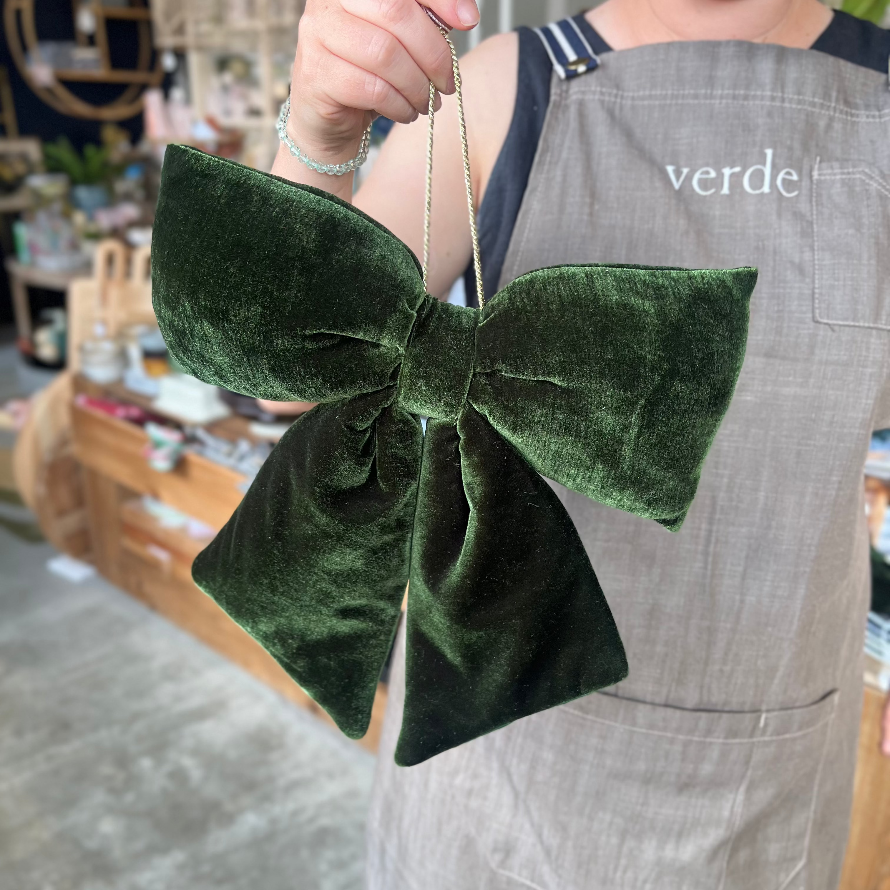 A person wearing a gray apron holds up a large dark green velvet bow in a shop setting.