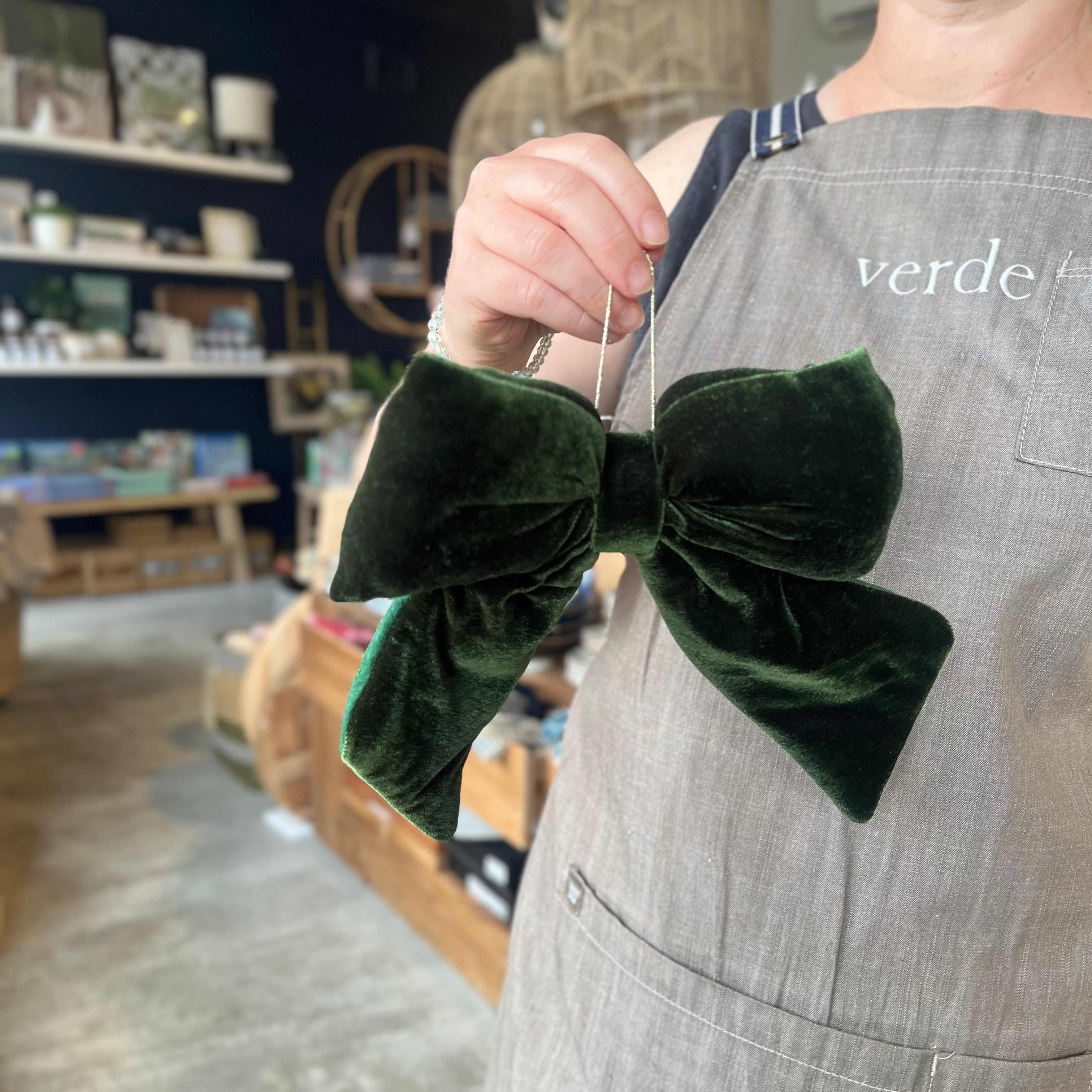 A person wearing a gray apron holds up a large, decorative dark green velvet bow in a shop setting.