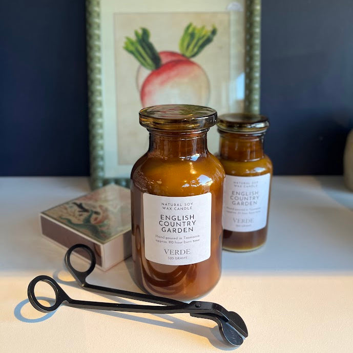 Two amber glass jars of scented candles with white labels reading 'English Country Garden' and 'Verde' are styled on a tabletop with a black metal candle wick cutter in front and a framed botanical print in the background.