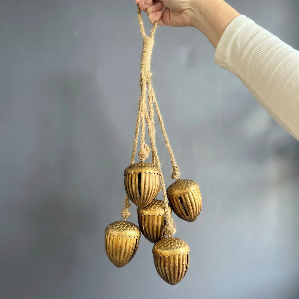 A hand wearing a cream ribbed long-sleeve shirt holds a decorative hanging set of five gold acorns suspended from a natural jute rope against a textured blue wall.