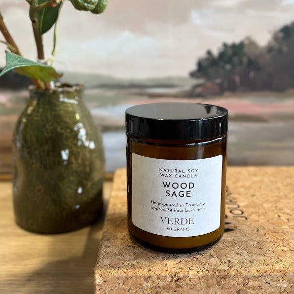 An amber jar candle labeled 'WOOD SAGE' by VERDE sits on a cork block on a wooden surface, with a vase of eucalyptus branches and a landscape painting in the background.