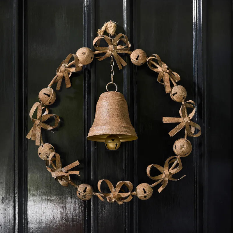 A rustic rusty wreath hanging on a black paneled door, featuring a central hanging bell and small jingle bells and bows around the circular garland.