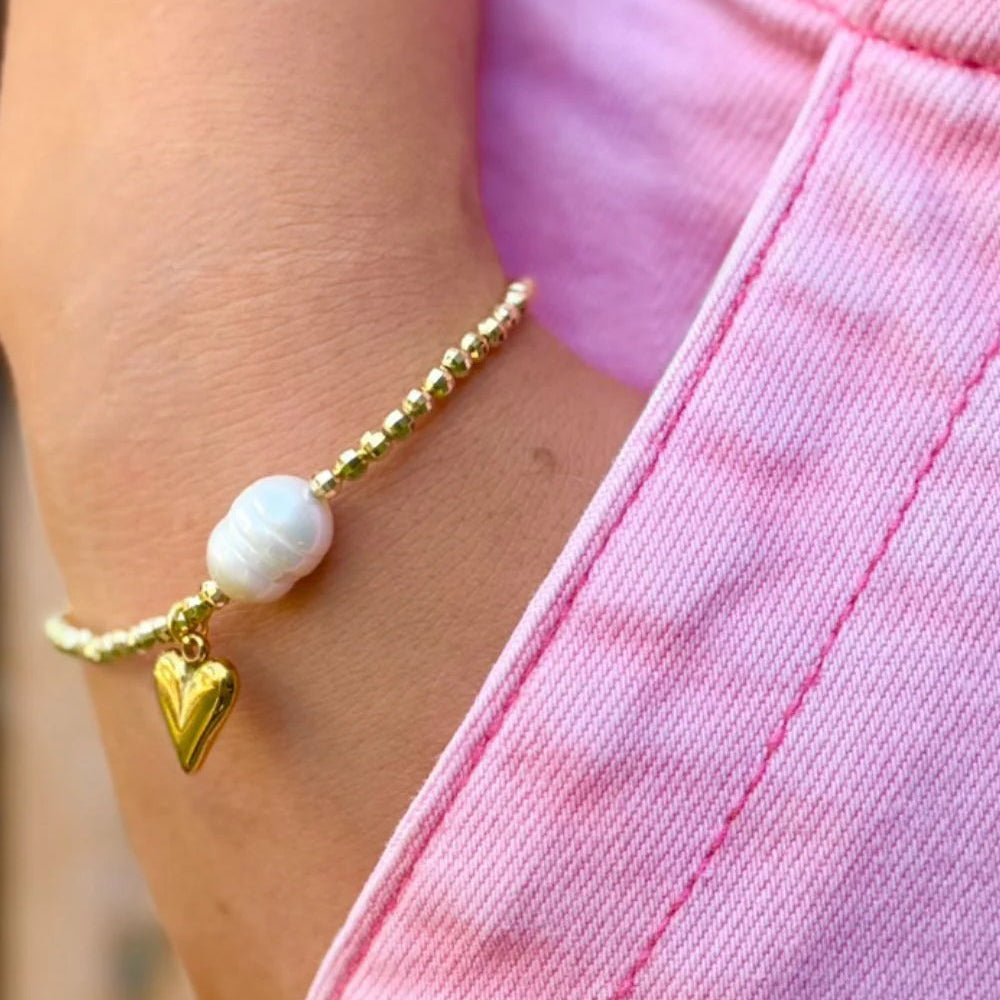 Gold bracelet with a heart charm and pearl bead on a person wearing a pink outfit.