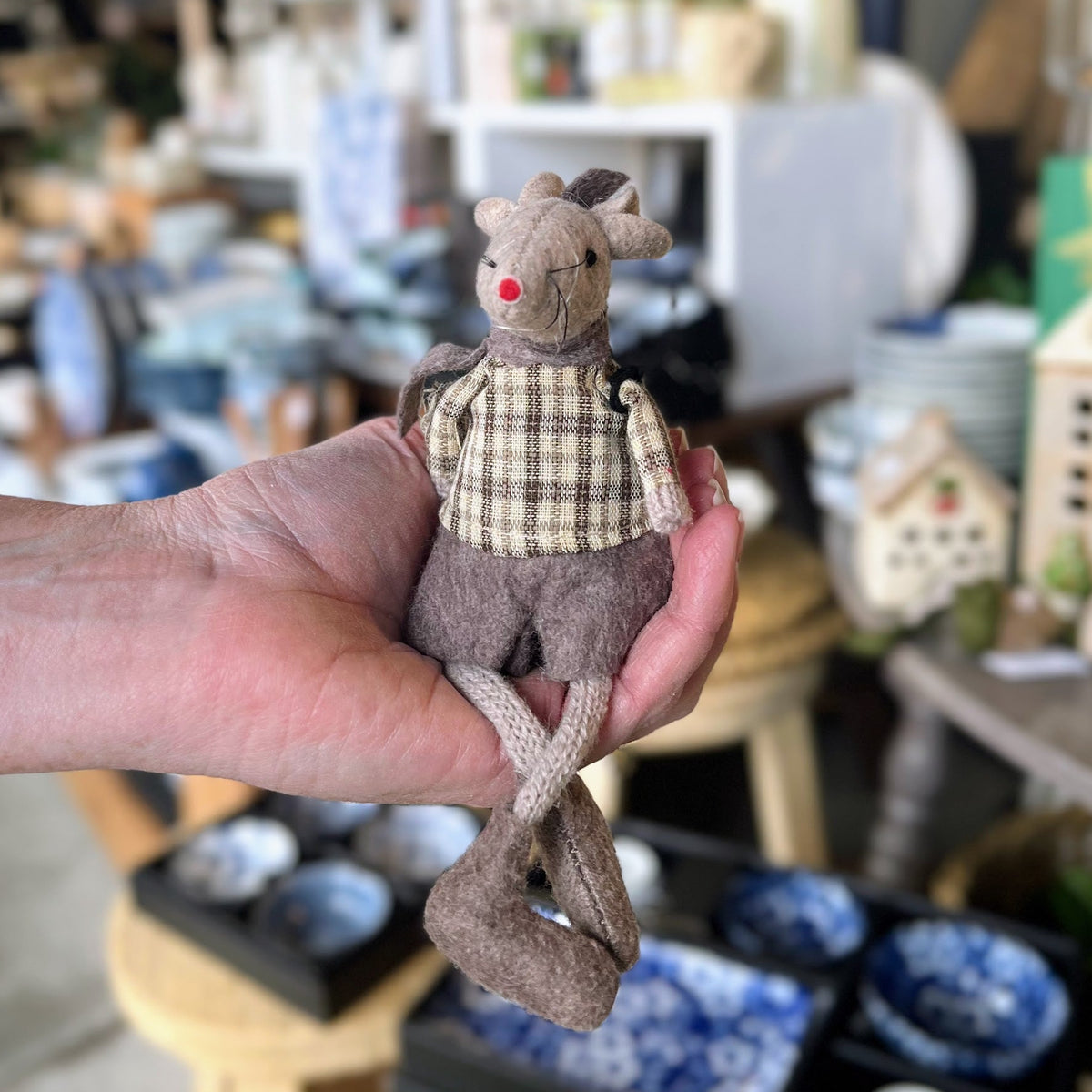 A hand holds a small plush toy mouse wearing a beige and brown plaid shirt and brown pants, with a blurred shop background filled with home decor items.
