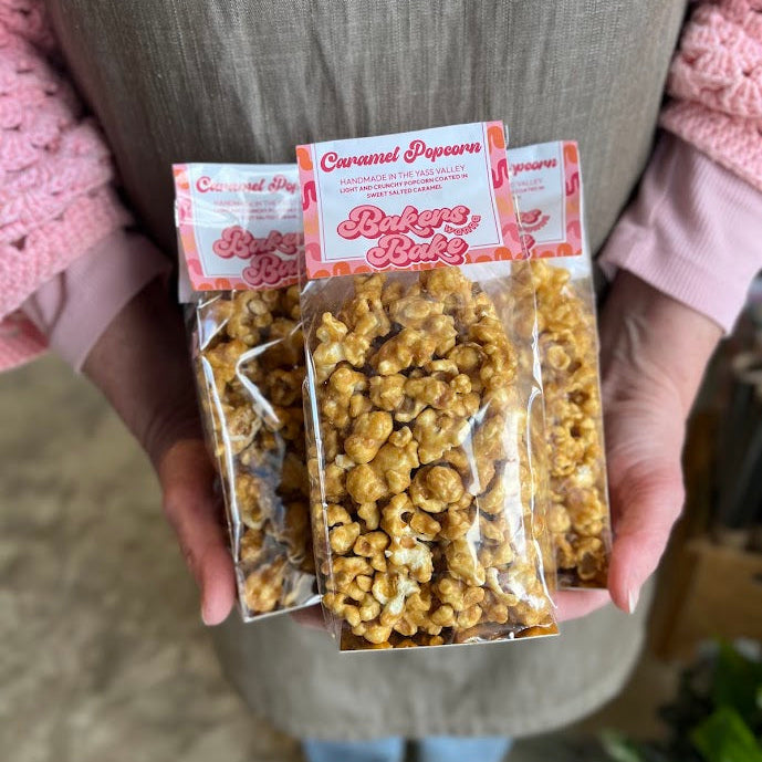 A person wearing a gray apron holds three clear bags of caramel popcorn. The bags have pink and white labels with the text 'Caramel Popcorn' 