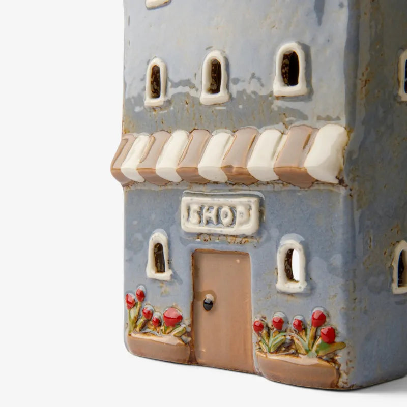 A close-up of a decorative ceramic miniature shop building with a distressed blue-gray facade, cream awnings, a brown door, and small red flower planters, shown against a plain white background.