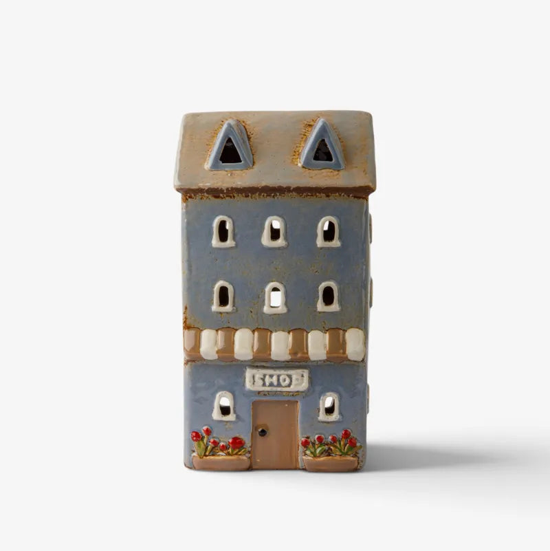 A small decorative building-shaped ceramic ornament or candle holder with a blue-gray facade, brown roof, arched windows, a door, and small red flower details, shown on a plain light background.