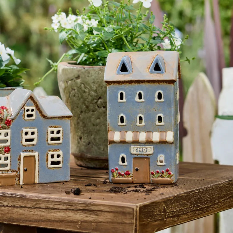 Two decorative miniature house-shaped garden planters sit on a rustic wooden table outdoors, with a blurred green garden and white flowers in the background.