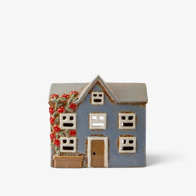 A small decorative ceramic house figurine with a blue-gray body, brown roof, white window frames, a brown door, and red flowers climbing up the left side, shown on a plain light background.