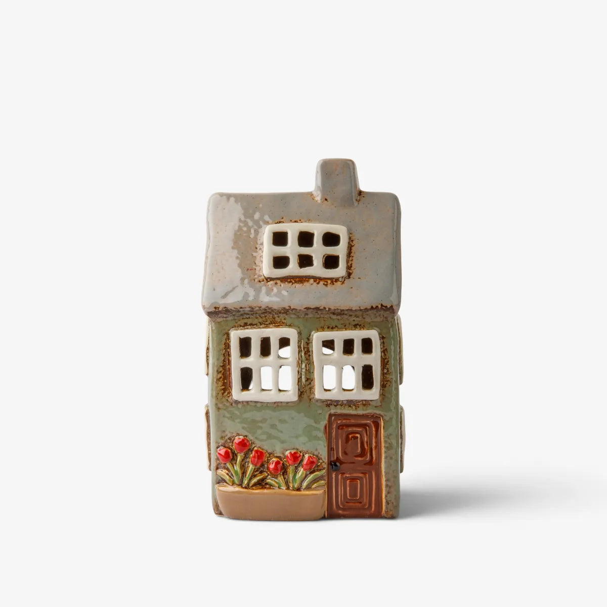 A small ceramic house-shaped candle holder or decorative figurine with a weathered green facade, brown door, white window panes, and red flowers at the base, shown on a plain light gray background.
