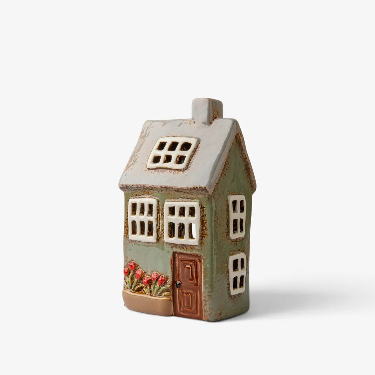 A small decorative ceramic house figurine with a distressed sage green body, white window panes, a brown door, and red flowers in a front planter, shown on a plain light gray background.