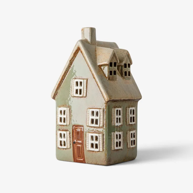 A small decorative ceramic house figurine with a distressed sage green and beige body, white window frames, and a brown door, shown on a light gray background.