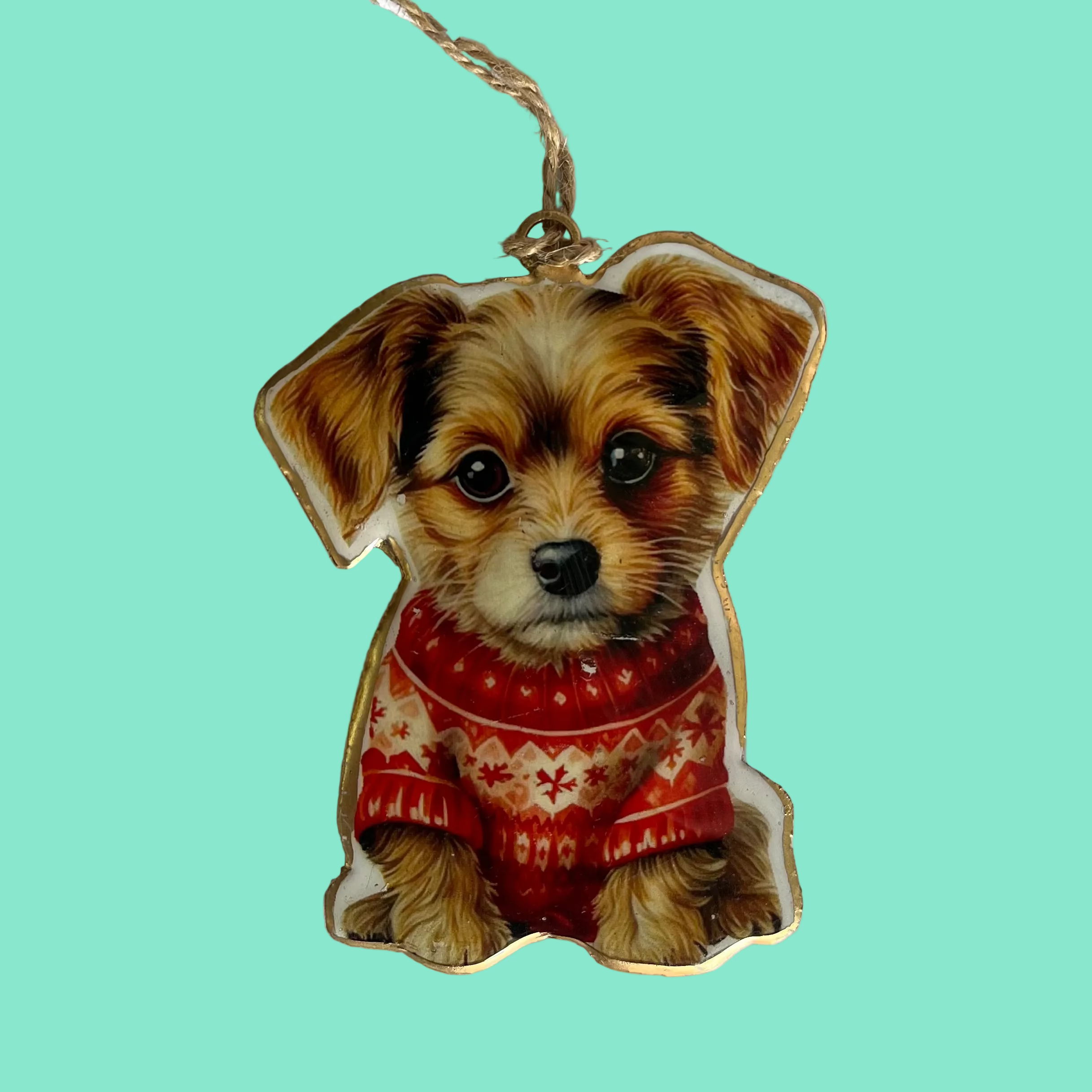 A \dog-shaped Christmas ornament featuring a printed photo of a small brown and white puppy wearing a red patterned sweater, hanging from a natural jute string on a light green background.
