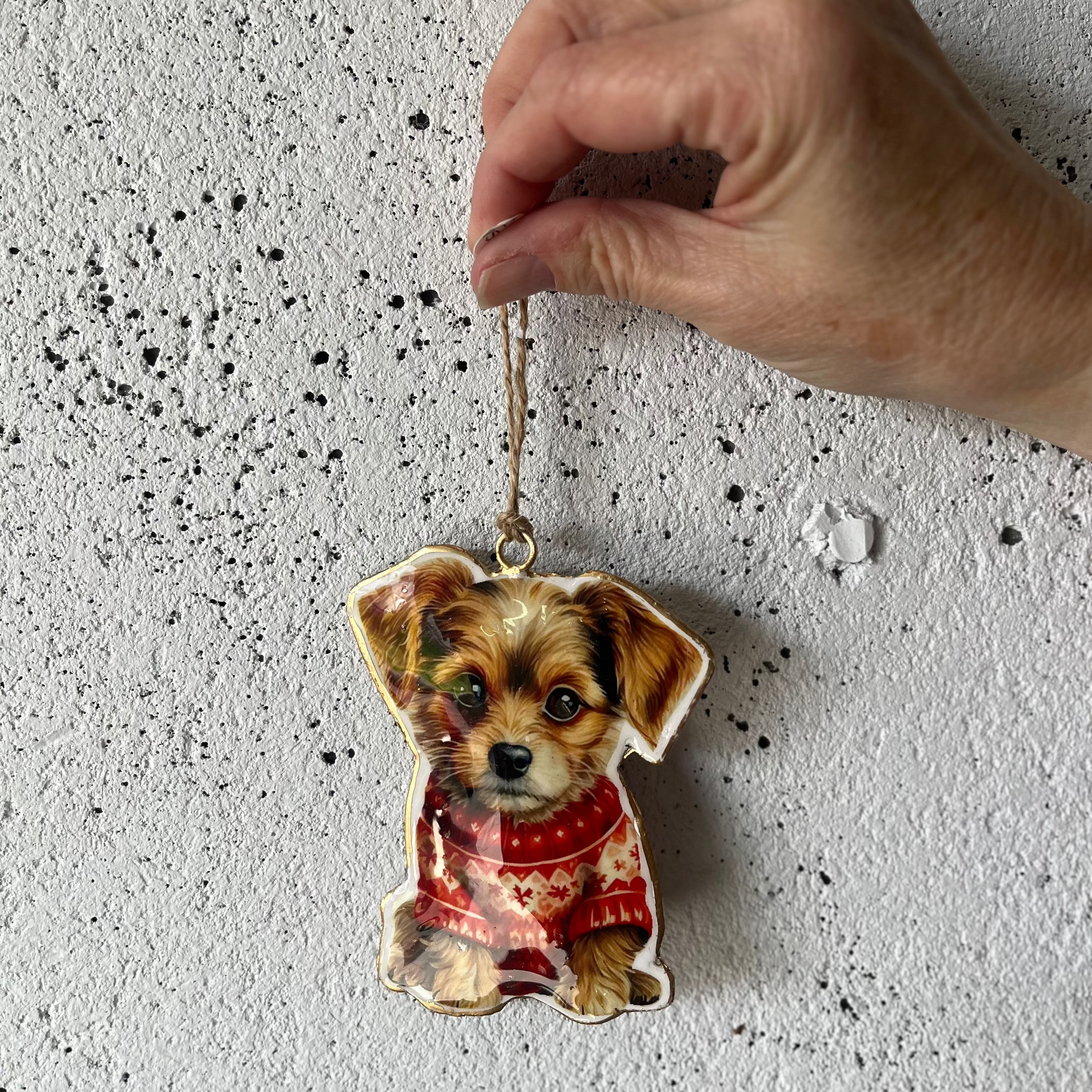 A hand holding a small dog-shaped ornament wearing a red and white patterned sweater, photographed against a textured light gray wall.