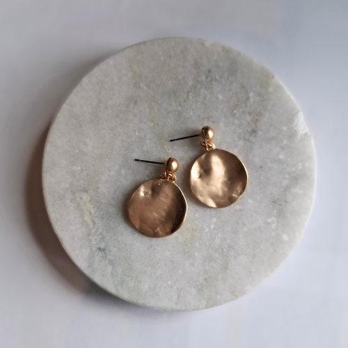 A pair of gold beaten disc earrings on a round marble surface