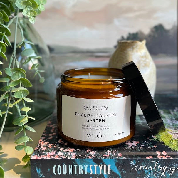 A jar candle labeled 'English Country Garden' by verde, placed on a decorative book with a black lid leaning against it, styled with greenery and a framed landscape print in the background.