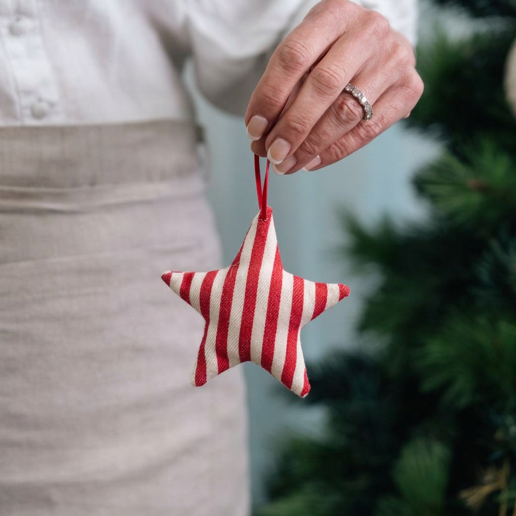 A hand holding a red and white striped fabric star ornament with a red hanging loop, shown against a soft-focus background with a person wearing an apron and green foliage on the right.