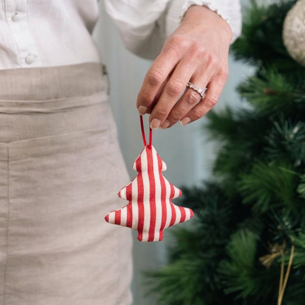 A person wearing a beige apron with holds a red and white striped fabric Christmas tree ornament in front of a blurred green Christmas tree.