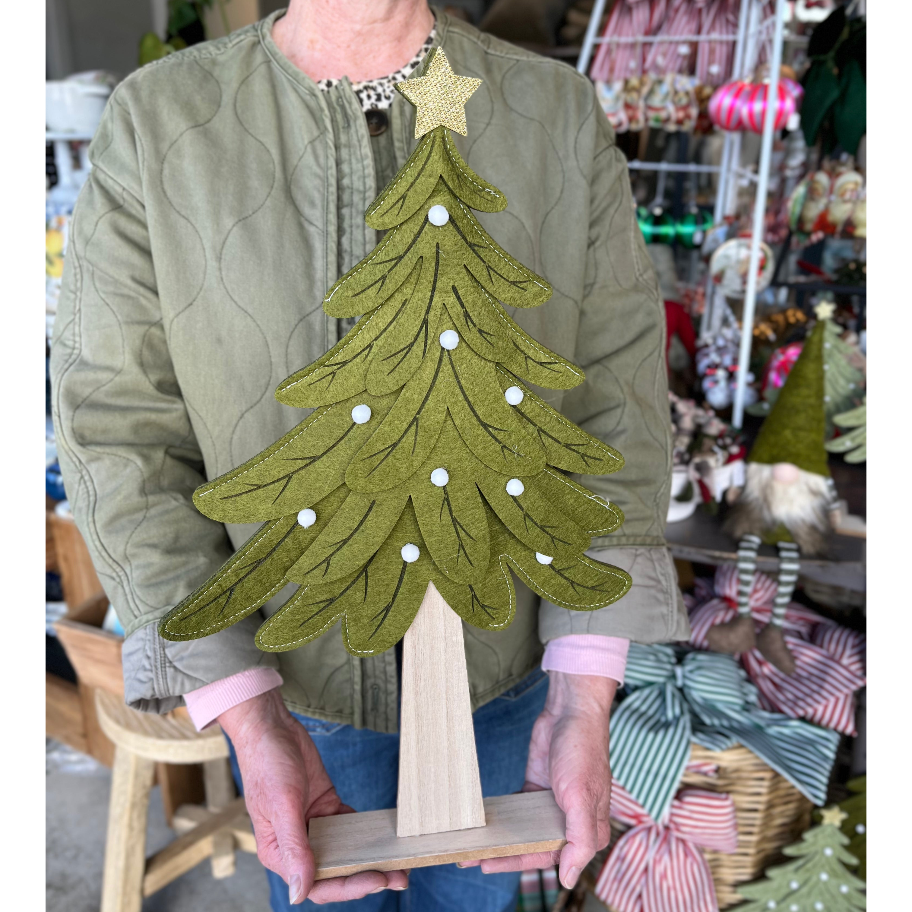 A person in a quilted olive green jacket holds a felt Christmas tree decoration with a gold star on a wooden stand in a craft shop.