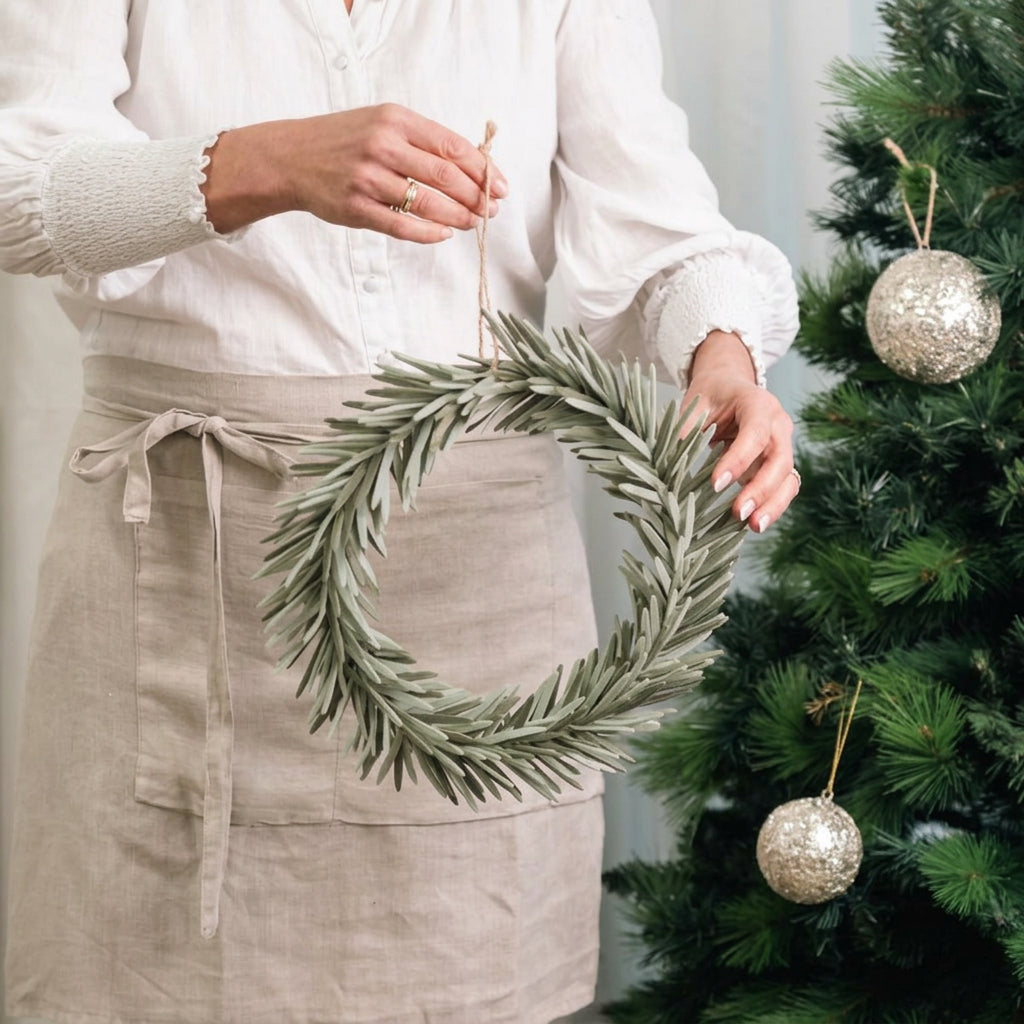 A person wearing a white blouse and a beige linen apron hangs a green pine wreath with silver glittery ornaments next to a decorated Christmas tree.